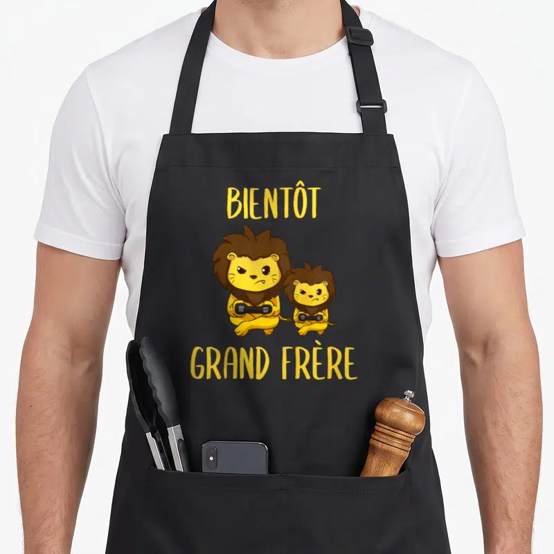 Tablier de cuisine noir avec deux lions jaunes tenant des manettes et le texte Bientôt Grand Frère en jaune.