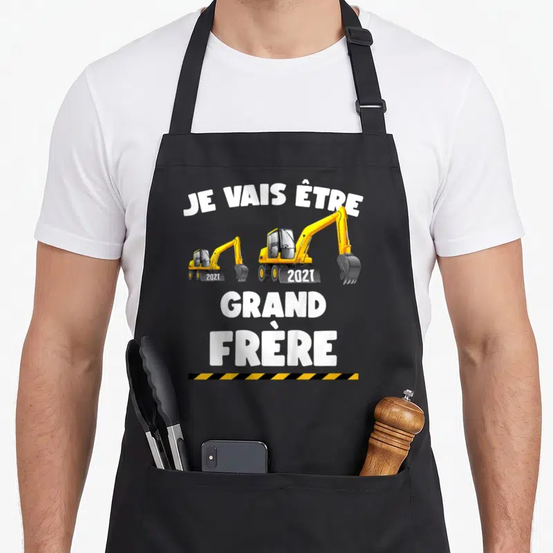 Tablier de cuisine noir avec deux excavatrices jaunes et le texte blanc Je vais être grand frère 2021 sur fond uni.