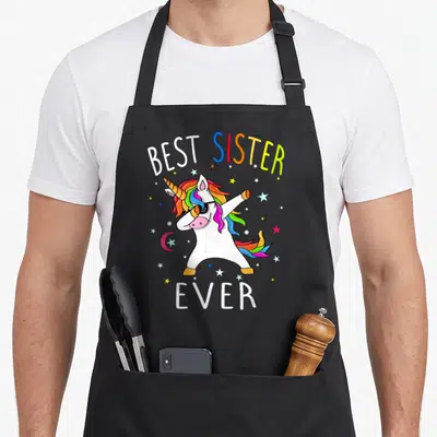 Tablier de cuisine noir avec une licorne blanche aux cheveux arc-en-ciel effectuant un dab sous des étoiles multicolores.
