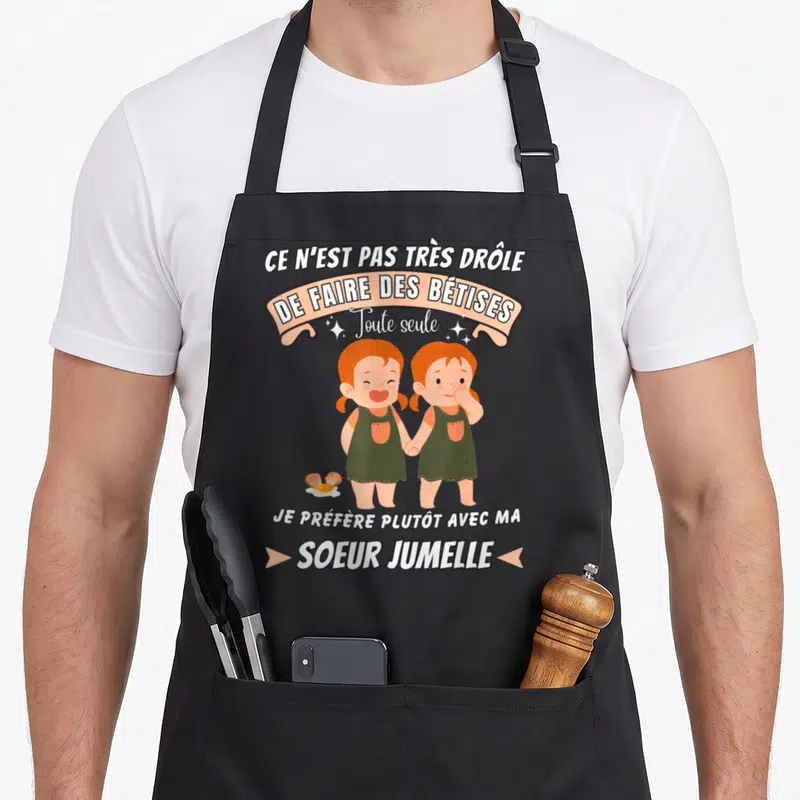 Tablier de cuisine noir avec illustration de deux filles jumelles aux cheveux roux et un œuf au sol, sous un bandeau rose.