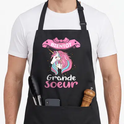 Tablier de cuisine noir avec deux licornes roses et le texte Bientôt grande soeur sous un ruban.
