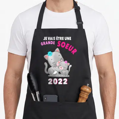 Tablier de cuisine noir avec deux chats gris s'embrassant et le texte Je vais être une grande soeur 2022 en rose et blanc.