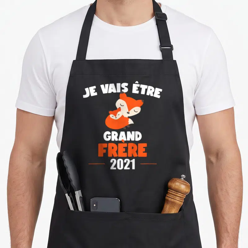 Tablier de cuisine noir avec deux renards orange et le texte Je vais être grand frère 2021 imprimé en blanc et orange.