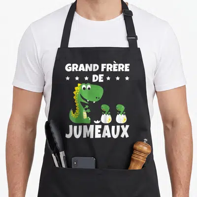 Tablier de cuisine noir avec un dinosaure vert et deux bébés dinosaures sortant d'œufs, texte blanc en haut et en bas.