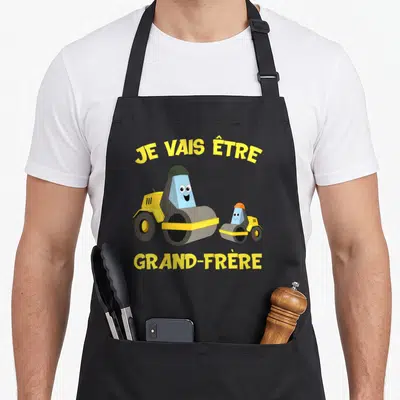 Tablier de cuisine noir avec deux rouleaux compresseurs jaunes et bleus et le texte Je vais être grand-frère en jaune.