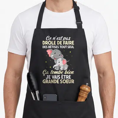Tablier de cuisine noir avec deux éléphants gris et roses et un message sur la venue d'une grande sœur.