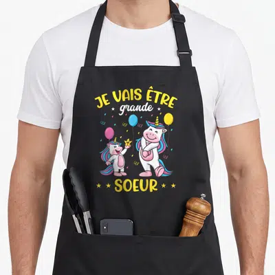 Tablier de cuisine noir avec deux licornes roses et des ballons sous le texte Je vais être grande soeur.