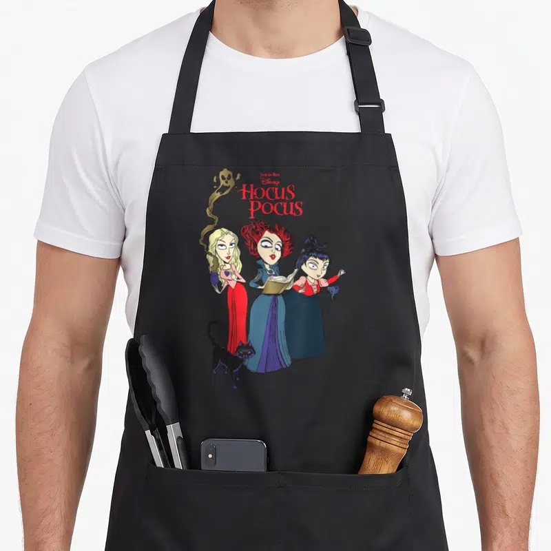 Tablier de cuisine noir avec illustration des trois sorcières Sanderson et un chat noir sous le titre Hocus Pocus en lettres rouges.