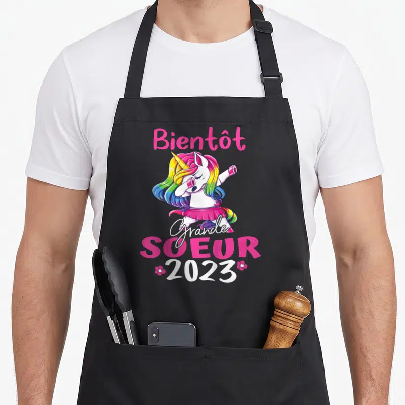 Tablier de cuisine noir avec licorne aux cheveux arc-en-ciel effectuant un dab et le texte Bientôt Grande Soeur 2023.