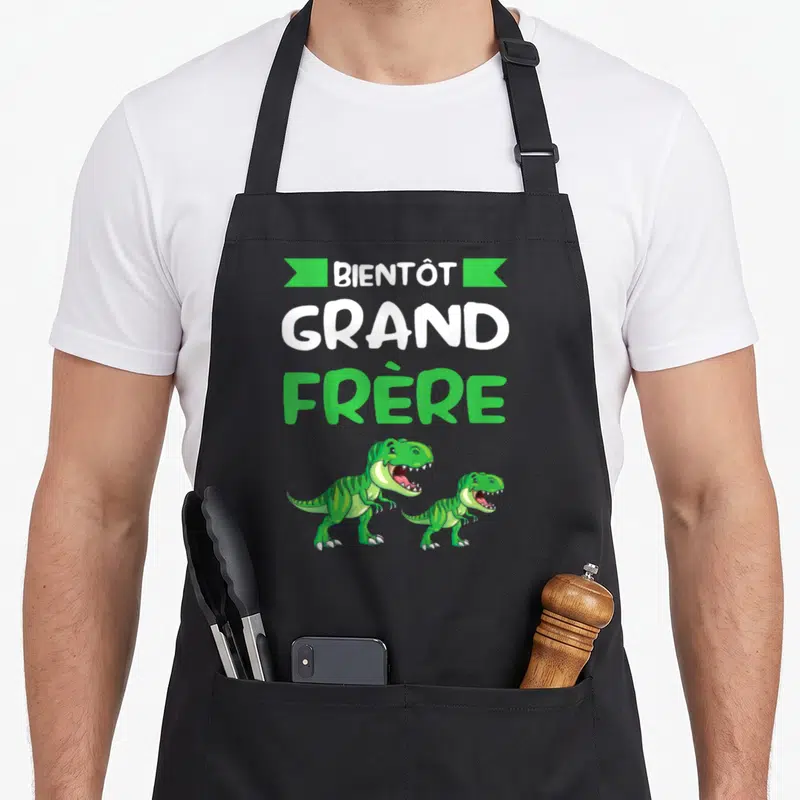 Tablier de cuisine noir avec inscription Bientôt Grand Frère et deux dinosaures Tyrannosaure verts en bas du motif.