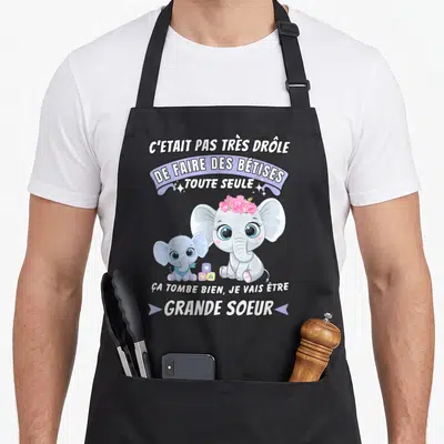 Tablier de cuisine noir avec deux éléphants roses et le texte Je vais être grande soeur 2024 imprimé en blanc.