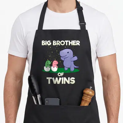 Tablier de cuisine noir avec illustration d'un dinosaure violet et deux bébés dinosaures vert et rose dans des coquilles d'œuf.