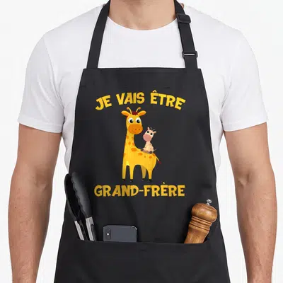 Tablier de cuisine noir avec une girafe jaune portant un bébé et le texte Je vais être grand-frère en lettres jaunes.