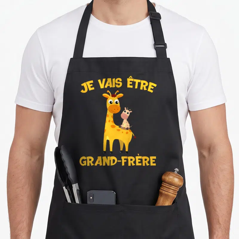 Tablier de cuisine noir avec une girafe jaune portant un bébé et le texte Je vais être grand-frère en lettres jaunes.