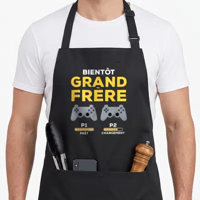 Tablier de cuisine noir avec texte Bientôt Grand Frère et deux manettes de jeu vidéo gris sur fond uni.