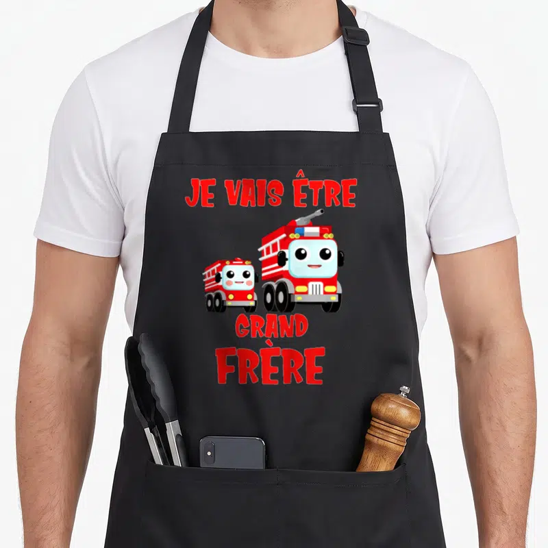 Tablier de cuisine noir avec deux camions de pompiers rouges et l'inscription Je vais être grand frère en lettres capitales.