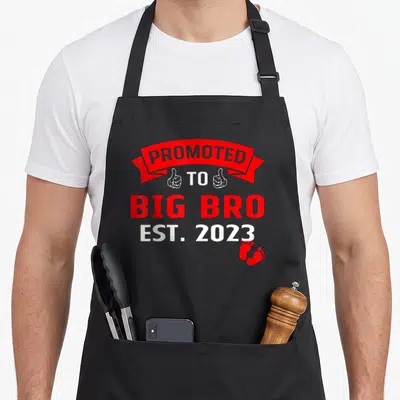 Tablier de cuisine noir avec motif « Promu Grand Frère » » » » 2023 en rouge et blanc incluant des pouces levés et des empreintes de pieds.