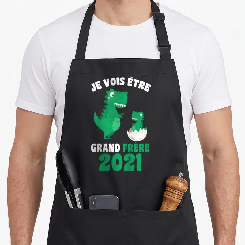 Tablier de cuisine noir avec deux dinosaures verts et l'inscription Je vois être grand frère 2021 en blanc et vert.