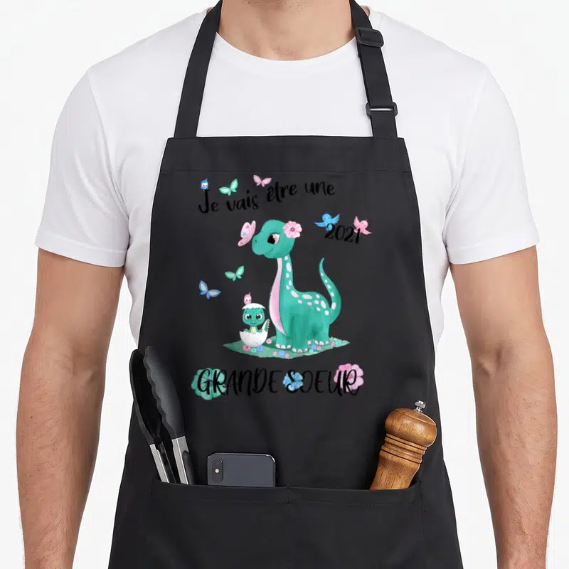 Tablier de cuisine noir avec un dinosaure vert, un bébé dino dans une coque et le texte Je vais être une grande soeur 2021.