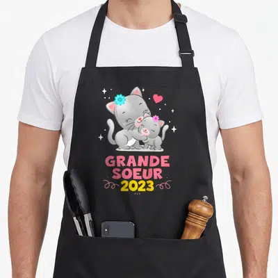Tablier de cuisine noir avec deux chats gris s'embrassant et le texte Grande Soeur 2023 en lettres colorées.