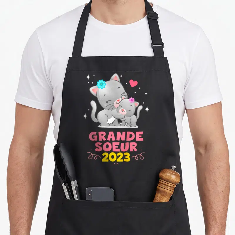 Tablier de cuisine noir avec deux chats gris s'embrassant et le texte Grande Soeur 2023 en lettres colorées.