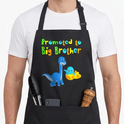 Tablier de cuisine noir avec illustration de dinosaures et texte « Promoted to Big Brother » en lettres colorées.
