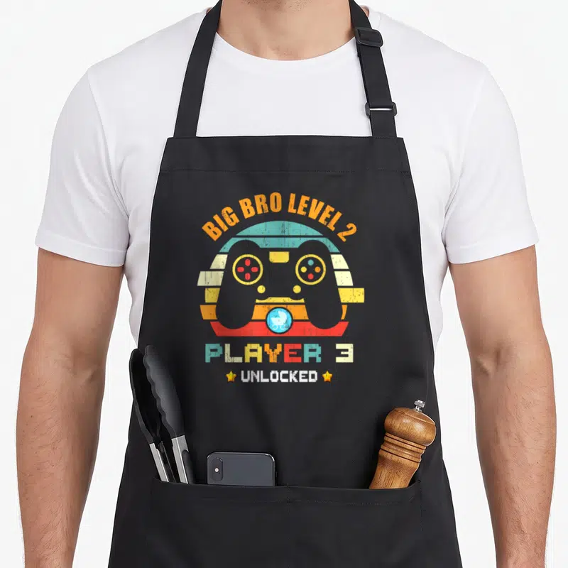Tablier de cuisine noir avec manette de jeu vidéo et texte Big Bro Level 2 Player 3 Unlocked en couleurs rétro.