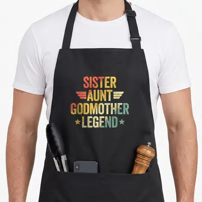 Tablier de cuisine noir avec texte Sister Aunt Godmother Legend coloré et motifs d'ailes stylisées.