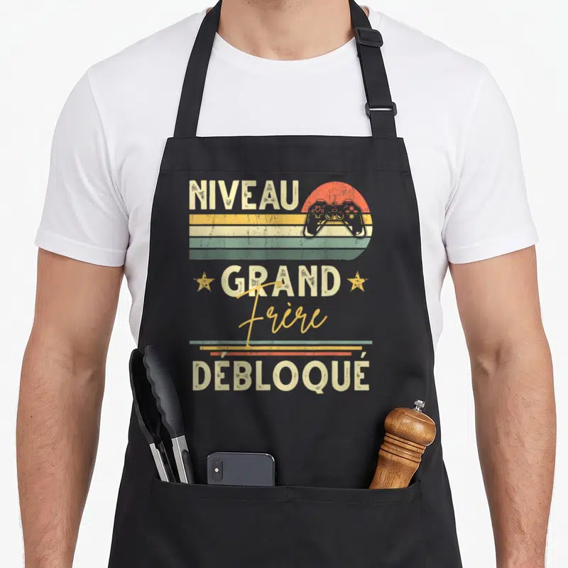 Tablier de cuisine noir avec illustration rétro Niveau Grand Frère Débloqué et manette de jeu vidéo.