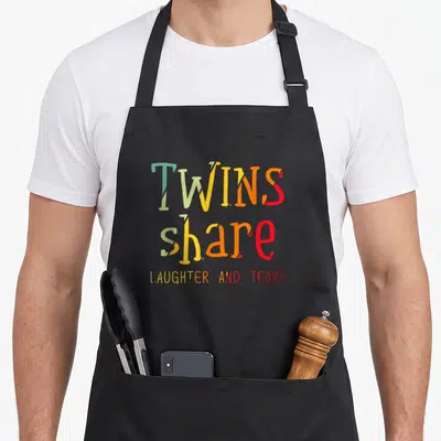 Tablier de cuisine noir avec inscription colorée Twins Share Laughter And Tears en typographie manuscrite.