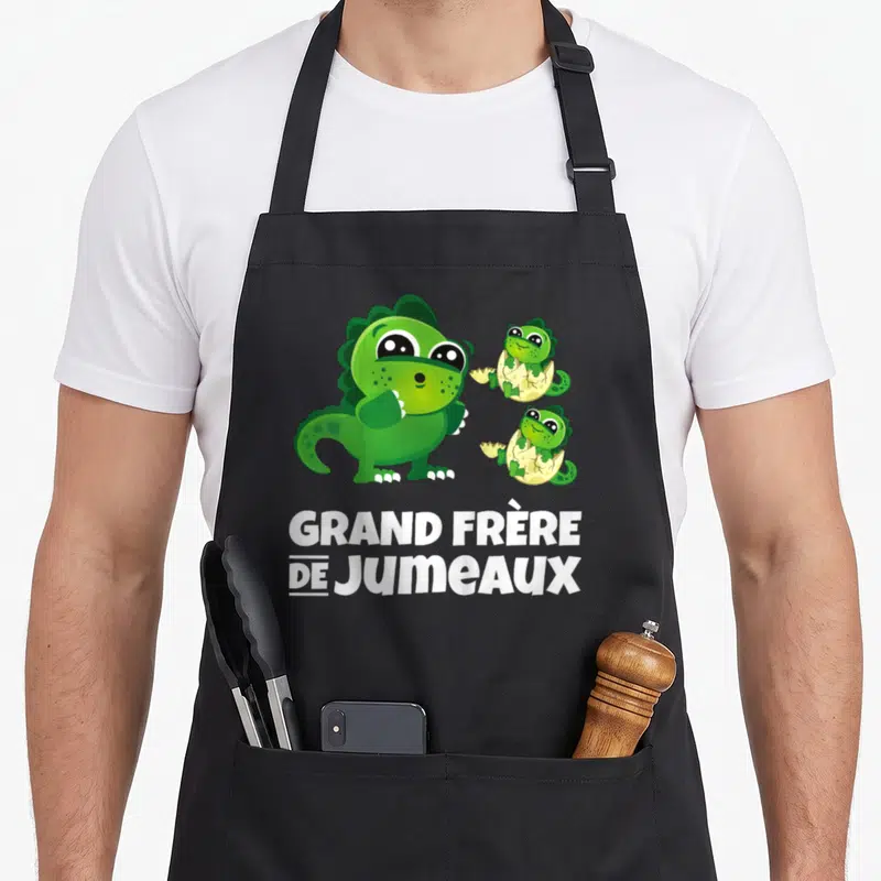 Tablier de cuisine noir avec trois dinosaures verts et l'inscription GRAND FRÈRE DE JUMEAUX en blanc.