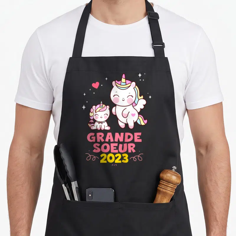 Tablier de cuisine noir avec deux licornes roses et l'inscription Grande Soeur 2023 en lettres vives.