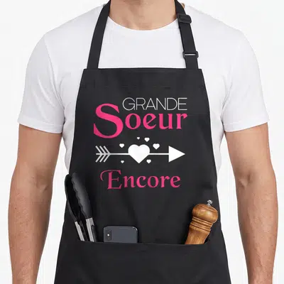 Tablier de cuisine noir avec inscription Grande Soeur Encore et une flèche traversant un cœur blanc.