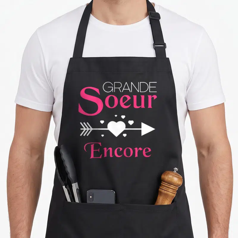 Tablier de cuisine noir avec inscription Grande Soeur Encore et une flèche traversant un cœur blanc.