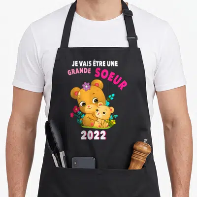 Tablier de cuisine noir avec ours marron et inscription Je Vais Être Une Grande Soeur 2022 entourés de fleurs roses et bleues.