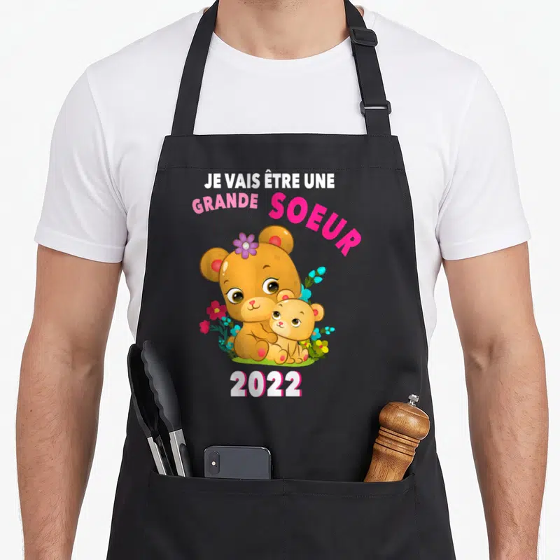 Tablier de cuisine noir avec ours marron et inscription Je Vais Être Une Grande Soeur 2022 entourés de fleurs roses et bleues.