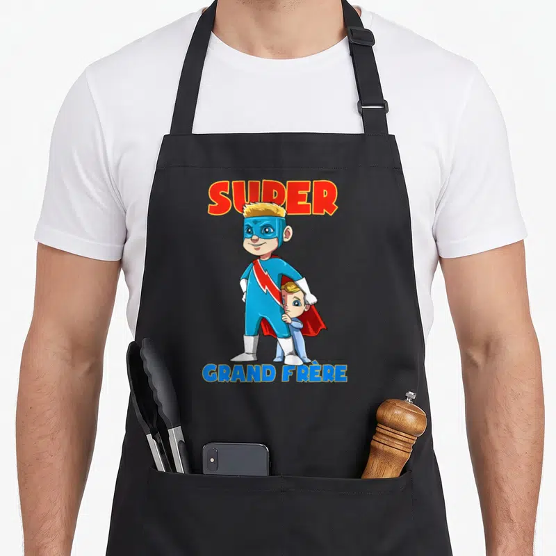Tablier de cuisine noir avec un super-héros blond et un enfant caché sous une cape rouge, texte SUPER GRAND FRÈRE en haut et en bas.