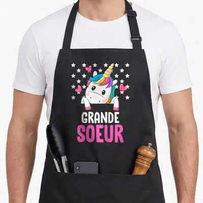 Tablier de cuisine noir avec une licorne rose et les mots Grande Soeur entourés d'étoiles et de cœurs.
