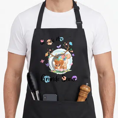 Tablier de cuisine noir avec un cerf floral et le texte Je vais être Grande Soeur 2027 imprimé au centre.