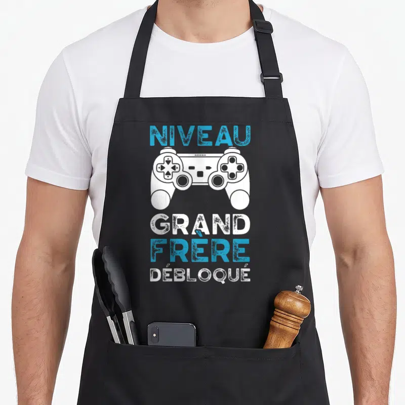 Tablier de cuisine noir avec manette blanche et texte Niveau Grand Frère Débloqué en bleu et blanc.