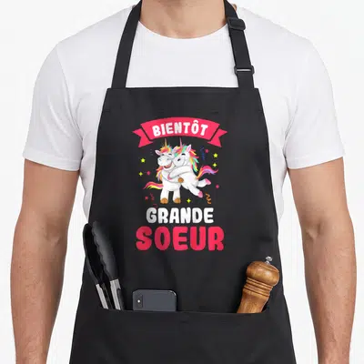Tablier de cuisine noir avec deux licornes s'embrassant et le texte Bientôt Grande Soeur en rose et blanc.