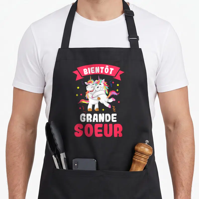 Tablier de cuisine noir avec deux licornes s'embrassant et le texte Bientôt Grande Soeur en rose et blanc.