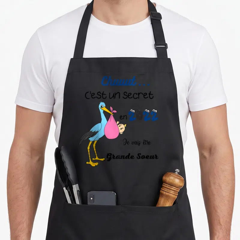 Tablier de cuisine noir avec aigle bleu portant un bébé dans un sac rose et texte announcing une future grande sœur pour l'année 2022.