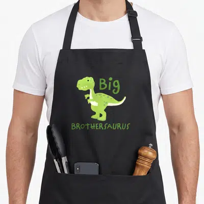 Tablier de cuisine noir avec dinosaure vert et texte Big Brothersaurus imprimé au centre.