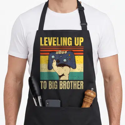 Tablier de cuisine noir avec illustration vintage main tenant une manette de jeu vidéo et texte leveling up to big brother en jaune et beige.