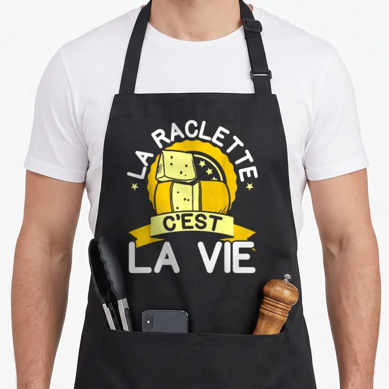 Tablier de cuisine noir avec une illustration colorée représentant des tranches de raclette et le texte La Raclette C'est La Vie.