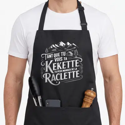 Tablier de cuisine noir avec inscription blanche Kekette Raclette et illustration de montagnes chalets et sapins.