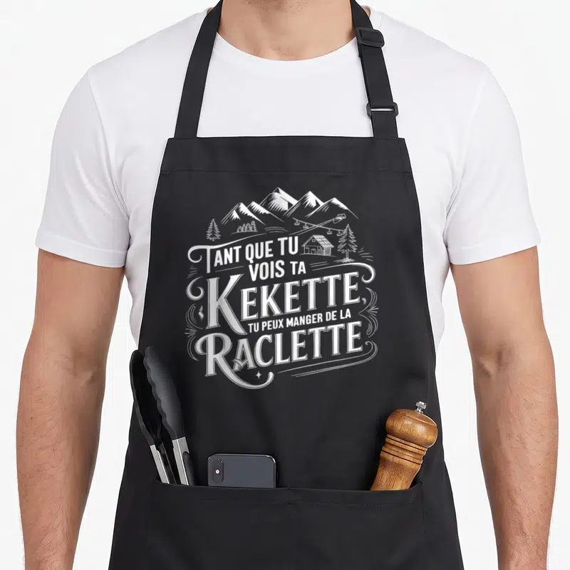 Tablier de cuisine noir avec inscription blanche Kekette Raclette et illustration de montagnes chalets et sapins.