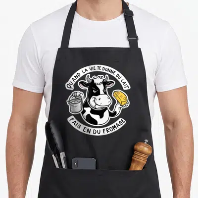 Tablier de cuisine noir avec une vache cartoon tenant un seau et un fromage jaune.