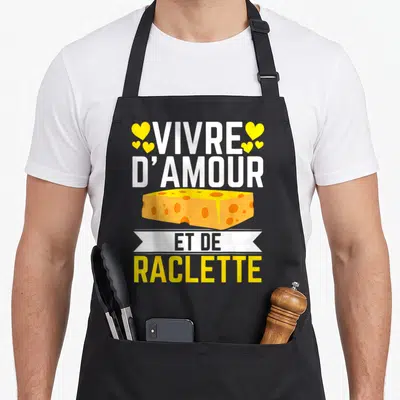 Tablier de cuisine noir avec illustration de bloc de fromage jaune, cœurs et texte Vivre d'amour et de raclette en blanc et jaune.